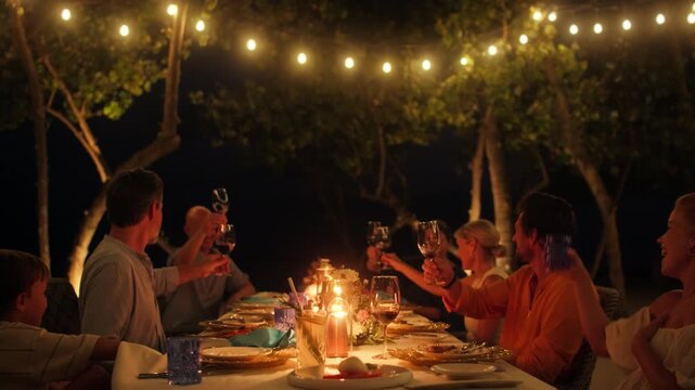 A multi-generational family and friends share a joyful dinner and toast outdoors under string lights at an island resort, celebrating connection and happiness together.
