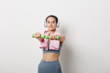 Young woman fitness portrait performing a front raise with green dumbbells, wearing headphones and a pink towel over shoulders, gray sports bra and leggings against a clean white background.