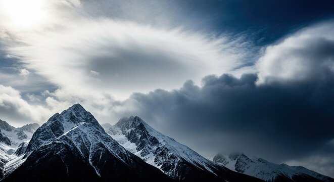 Dramatic snow capped mountain peaks pierce moody, dynamic clouds under bright sunlight creating an awe inspiring, majestic natural landscape.