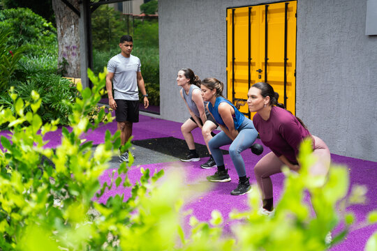 Latin hispanic fitness instructor guiding a diverse group of women performing kettlebell squats during an outdoor workout session