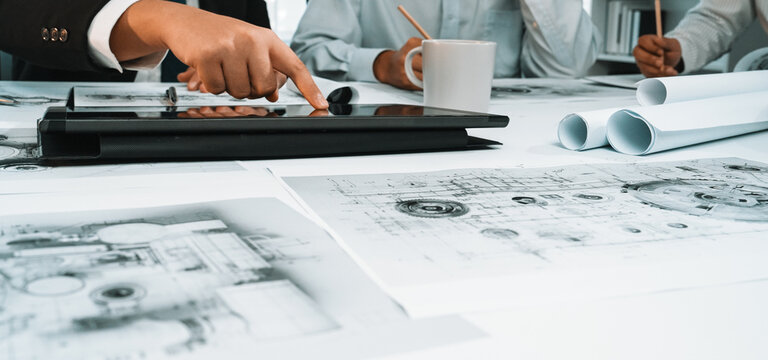 A diverse business team engages in an architectural project, utilizing a digital tablet while reviewing detailed blueprints and designs in a modern office setting. SACTR