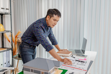A young architect works with building plans and a house model in the office, reviewing blueprints, marking details, and analyzing design structures with focus and creativity.