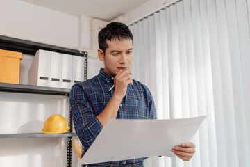 A young architect works with building plans and a house model in the office, reviewing blueprints, marking details, and analyzing design structures with focus and creativity.