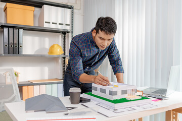 A young architect works with building plans and a house model in the office, reviewing blueprints, marking details, and analyzing design structures with focus and creativity.