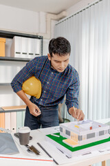 A young architect works with building plans and a house model in the office, reviewing blueprints, marking details, and analyzing design structures with focus and creativity.