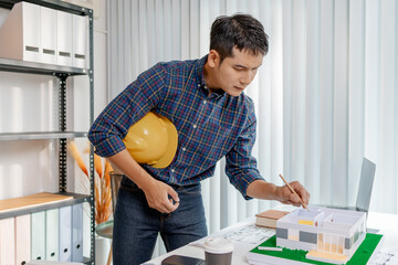 A young architect works with building plans and a house model in the office, reviewing blueprints, marking details, and analyzing design structures with focus and creativity.