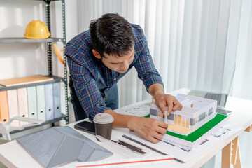 A young architect works with building plans and a house model in the office, reviewing blueprints, marking details, and analyzing design structures with focus and creativity.