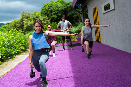 Athletic latin american women and men performing lunges with kettlebell weights during an outdoor fitness workout at modern gym - Powered by Adobe