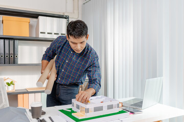 A young architect is working at his desk, reviewing blueprints, using a calculator, and checking a house model. He focuses on precision and detail while planning construction projects.