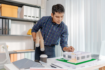 A young architect is working at his desk, reviewing blueprints, using a calculator, and checking a house model. He focuses on precision and detail while planning construction projects.