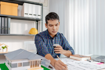 A young architect is working at his desk, reviewing blueprints, using a calculator, and checking a house model. He focuses on precision and detail while planning construction projects.