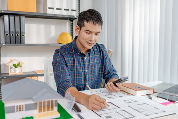 A young architect is working at his desk, reviewing blueprints, using a calculator, and checking a house model. He focuses on precision and detail while planning construction projects.