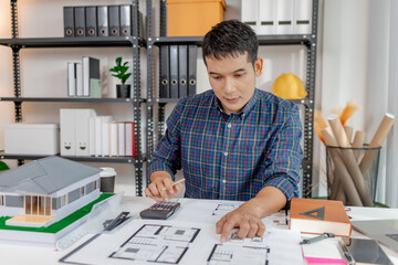 A young architect is working at his desk, reviewing blueprints, using a calculator, and checking a house model. He focuses on precision and detail while planning construction projects.