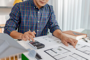 A young architect is working at his desk, reviewing blueprints, using a calculator, and checking a house model. He focuses on precision and detail while planning construction projects.