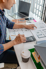 A young architect is working at his desk, reviewing blueprints, using a calculator, and checking a house model. He focuses on precision and detail while planning construction projects.