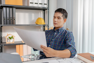 A young architect is working at his desk, reviewing blueprints, using a calculator, and checking a house model. He focuses on precision and detail while planning construction projects.