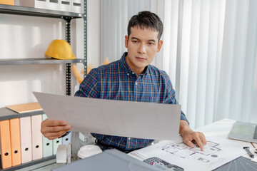 A young architect is working at his desk, reviewing blueprints, using a calculator, and checking a house model. He focuses on precision and detail while planning construction projects.