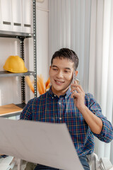 A young architect is standing at his desk while discussing a project on the phone, carefully reviewing construction plans with focus and attention in a modern office environment.
