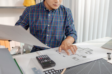 A young architect is working at his desk, reviewing blueprints, using a calculator, and checking a house model. He focuses on precision and detail while planning construction projects.