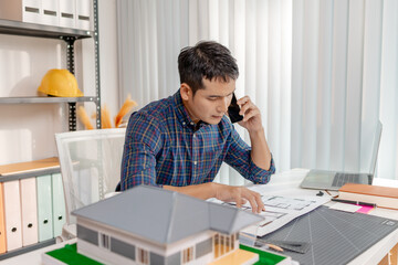 A young architect is standing at his desk while discussing a project on the phone, carefully reviewing construction plans with focus and attention in a modern office environment.