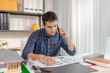 A young architect is standing at his desk while discussing a project on the phone, carefully reviewing construction plans with focus and attention in a modern office environment.