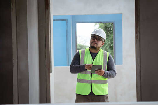 Asian overweight construction project foreman wearing white hard hat and reflective safety vest stands inspecting a construction project while holding a tablet and building plan in his hand.