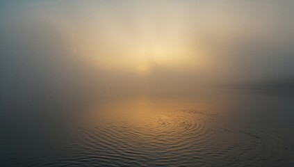 Obraz premium Water surface reflects sunlight through fog creating a hazy atmospheric effect.