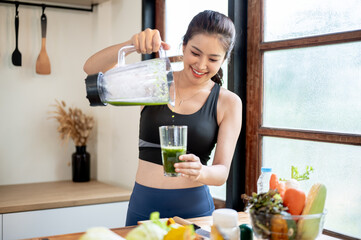Asian woman in sportswear pouring vegetable juice in blender to a glass standing at kitchen counter.