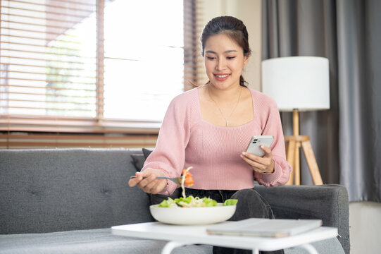 Asian woman holding phone and using fork eating vegetable salad while sitting on sofa in living room