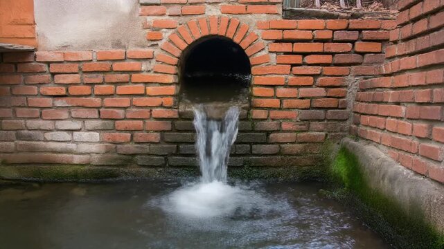 Clear water flows steadily from a traditional arched opening in a red brick wall into a pool below. Peaceful village drainage scene.