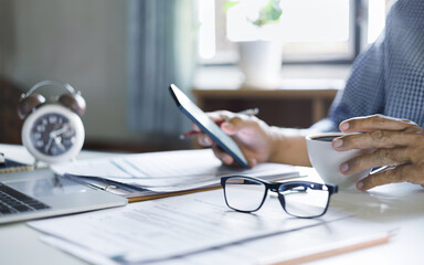 asian businessman working at home using mobile phone he taking a break during work in afternoon with coffee drink, select focus on hand holding coffee cup