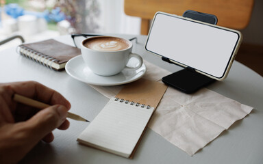 smart phone on stand with latte coffee, note book, diary and man hand holding pencile to taking note