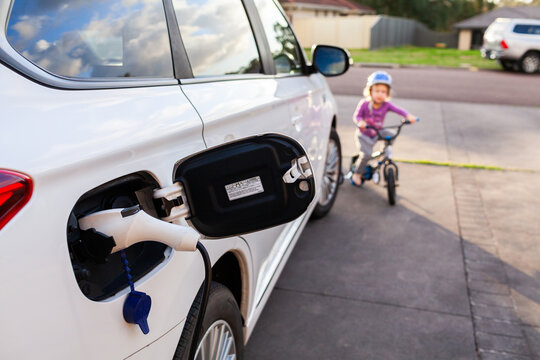 Electric vehicle charging in family home driveway with child riding bike in background