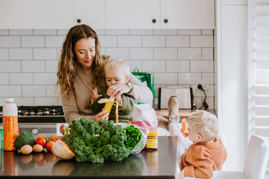 Mother peeling a banana for toddler son seated on countertop while other son eating a carrot