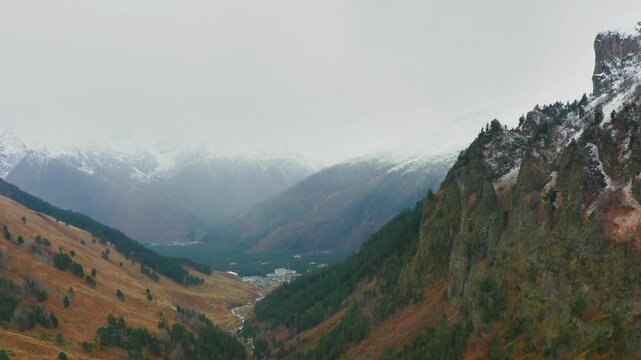 An aerial view of a scenic mountain valley in the North Caucasus region of Russia during autumn. The landscape features a river flowing through the valley, surrounded by mountains with patches of snow