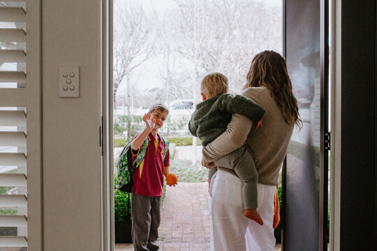 Mother holding toddler son in her hip saying goodbye to older son with backpack out the doorway