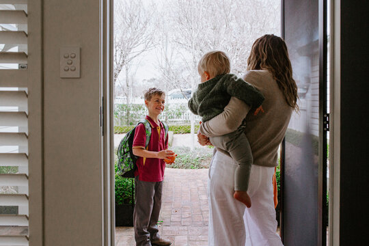 Mother holding toddler son in her hip saying goodbye to older son with backpack out the doorway