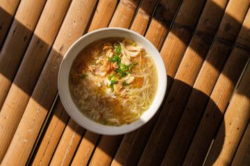Overhead shot of nusantara indonesian food calles soto filled with chicken noodle soup in a white bowl, placed on a bamboo mat with a shadow.