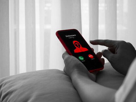 Scammer or fraud call alert concept with unknown caller appearing, red hooded figure on smartphone screen in hands, black and white photo. Phone phishing, cybercrime, and mobile security risk.