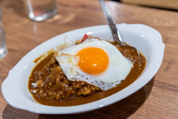 Close-up Japanese curry rice, Kare Raisu topped with a perfect sunny-side-up fried egg, served in a white bowl on a wooden table.