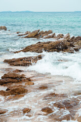 Waves crash on brown rock formation with white foam and bubble on coastal rocky sea beach with deep blue turquoise ocean, sky, islands in horizon in background for dynamic, dramatic nature backgroud
