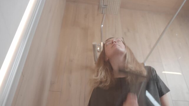Close-up of woman using rubber squeegee to clean glass shower door in modern bathroom. Concept of hygiene, home care and daily cleaning routine