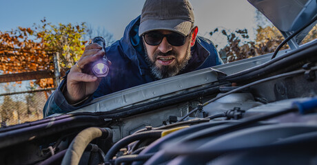 Man inspecting car engine with flashlight during early evening