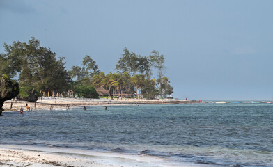 Watamu Beach, Kenya, during low tide with people playing and swimming in the shallow turquoise waters. The scene captures a lively coastal moment with small boats anchored in the background.