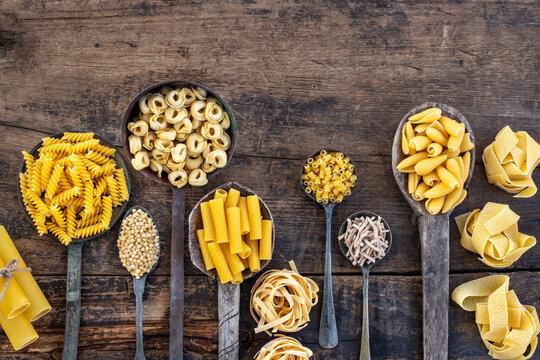 Different types of uncooked pasta on rustic wooden table, in the spoons, cutting board and bowls 