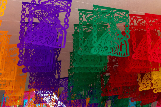 A colorful string of paper lanterns hang from the ceiling. The lanterns are in various colors. Mexican Day of the Dead tradition, offerings, food, colors, decorations, and papel picado, La Catrina.