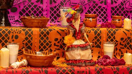 A table with a woman in a red dress and a flower on her head. There are also candles. Mexican Day of the Dead tradition, offerings, food, colors, decorations, and papel picado, La Catrina.