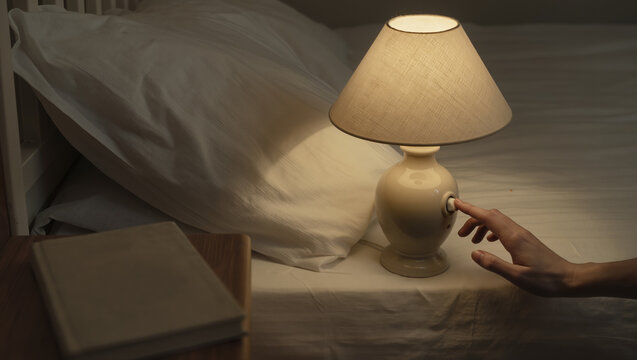 Cozy evening scene with a ceramic table lamp on a bed, warm light glowing, hand reaching to switch it off, and a closed book, evoking intimate reading moments