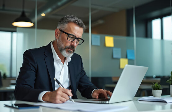 Mature businessman intently reviews paperwork at his desk in modern office. He signs documents and analyzes financial reports on a laptop. Focused pro works diligently with business strategy.