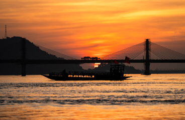 Obraz premium A boat sails on the river Brahmaputra during sunset, with the silhouette of a cable-stayed bridge and hills in the background